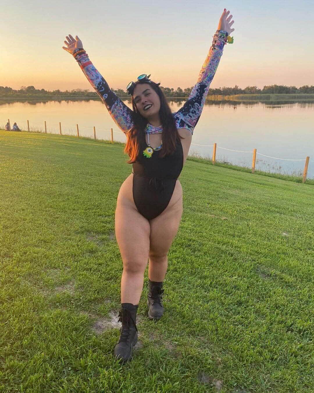 Woman wearing matte black rave bodysuit with colorful arm sleeves celebrating at sunset festival field