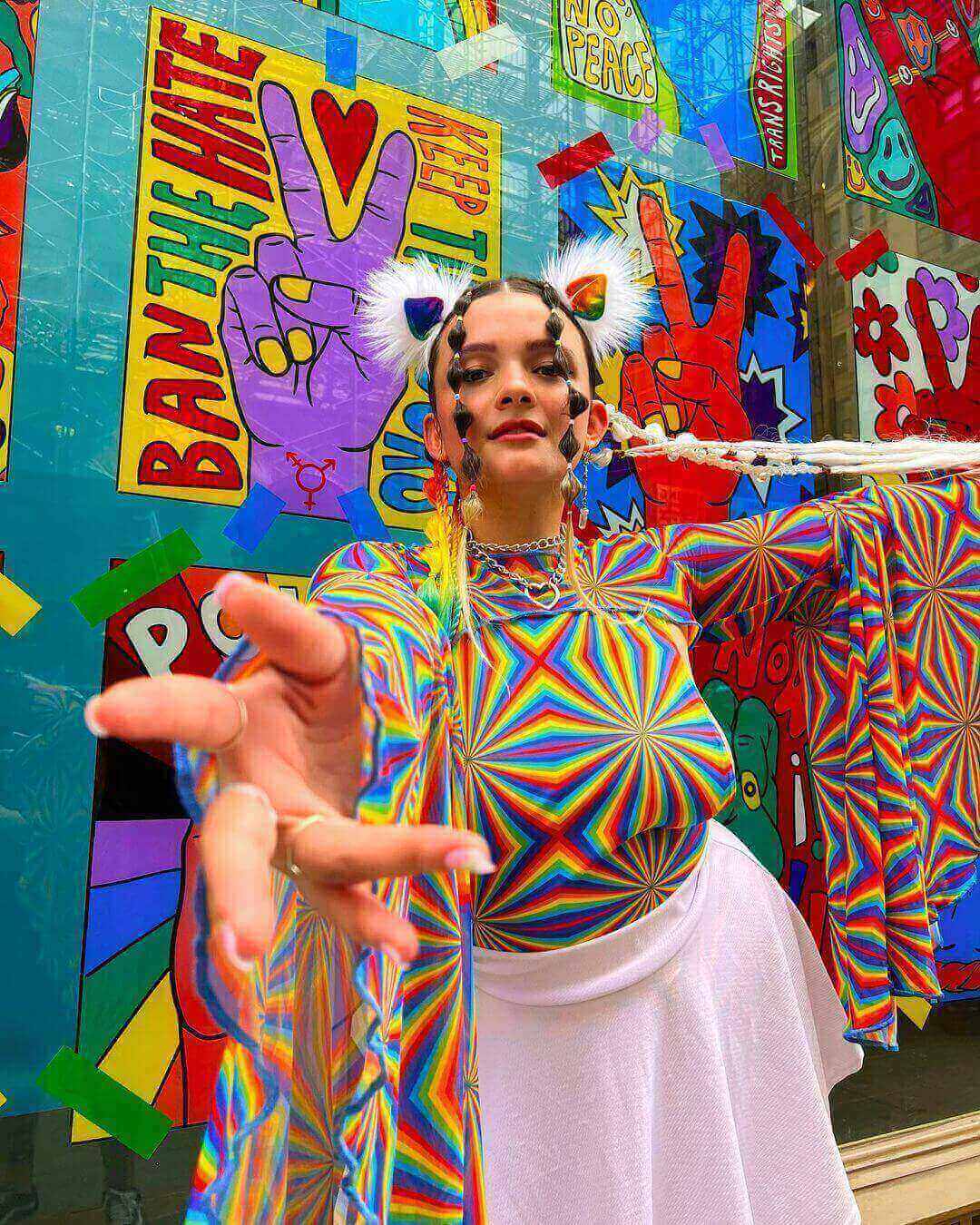 Model in white mesh rave skirt and rainbow top making peace sign against colorful festival graffiti wall