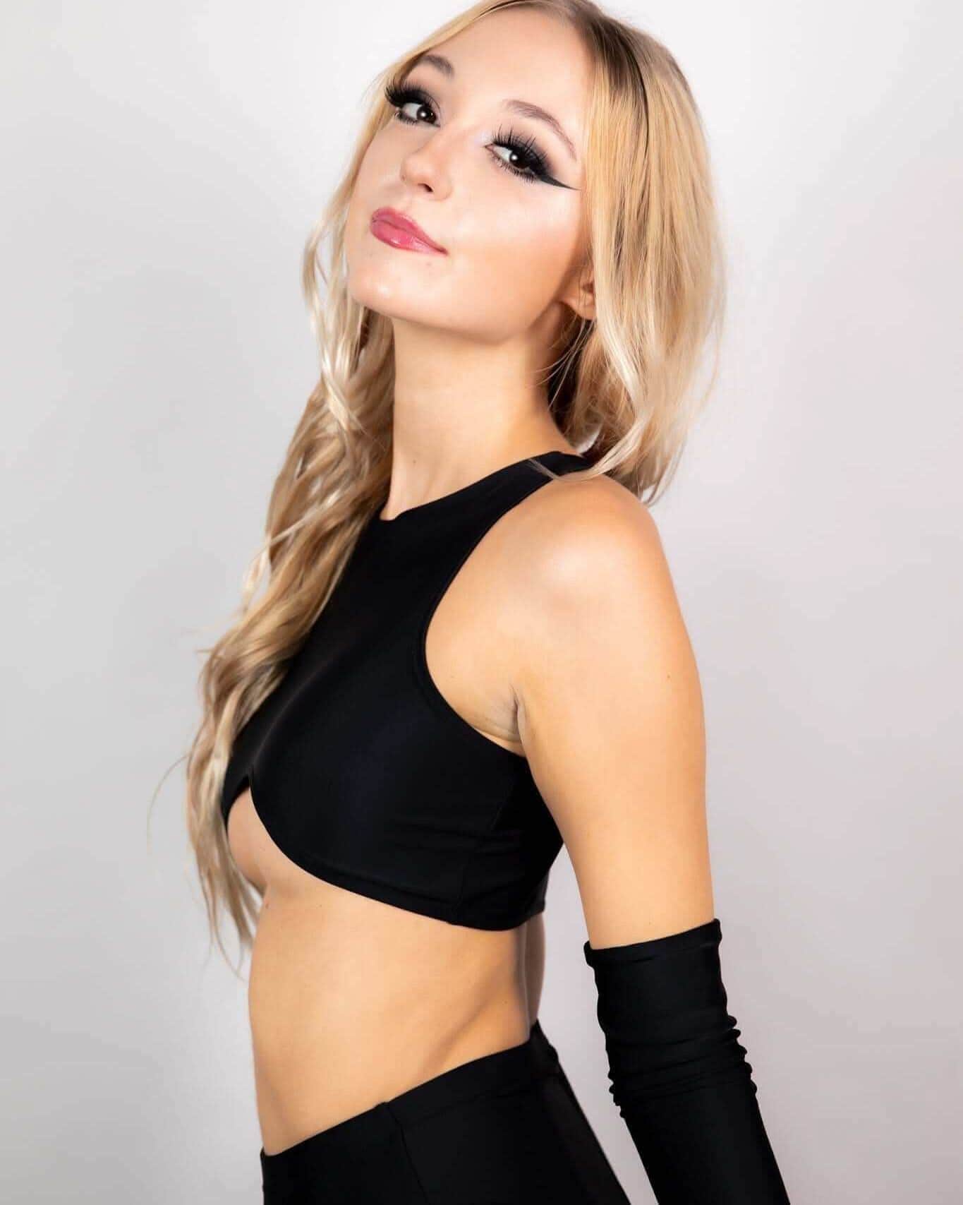 Blonde model wearing matte black teaser rave top with curved underboob cutout, side pose festival outfit styling