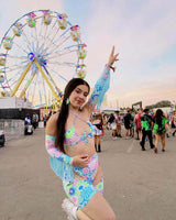 Young woman wearing pastel floral chaps and a festival outfit poses cheerfully by a ferris wheel at a rave event.