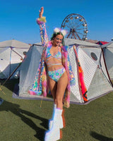 Young woman in pastel rave outfit with floral print, standing in front of tents and a Ferris wheel at a festival.