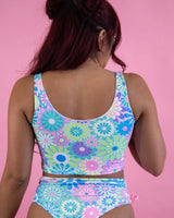 Model showcasing a pastel groovy flower print bikini top and bottom against a pink background, perfect for rave outfits.