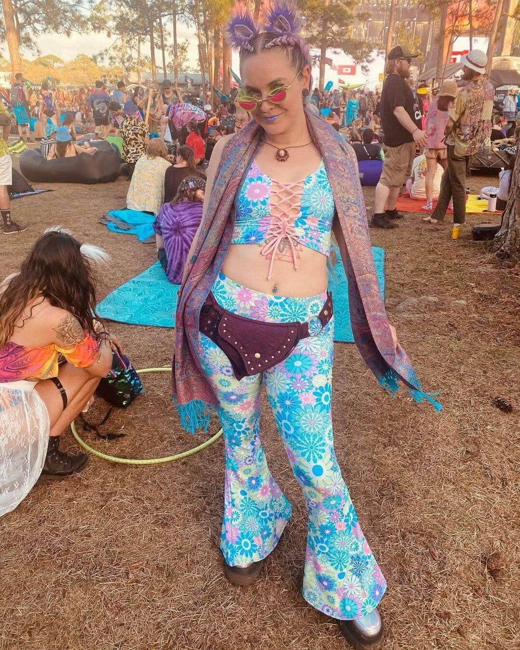 Woman in pastel groovy flower print lace-up tank top and flared pants at a vibrant outdoor rave festival.