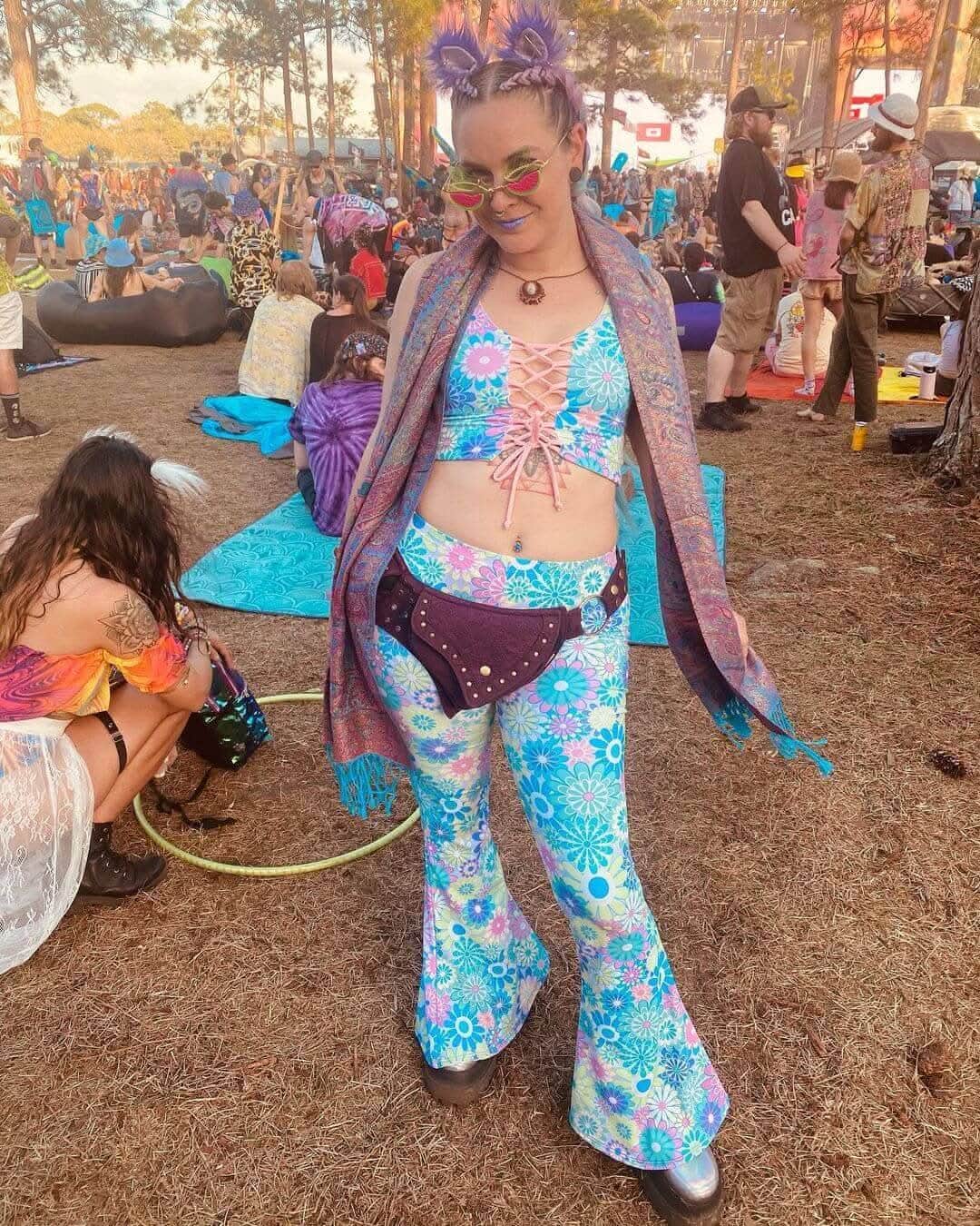 Woman in pastel groovy flower print lace-up tank top and flared pants at a vibrant outdoor rave festival.