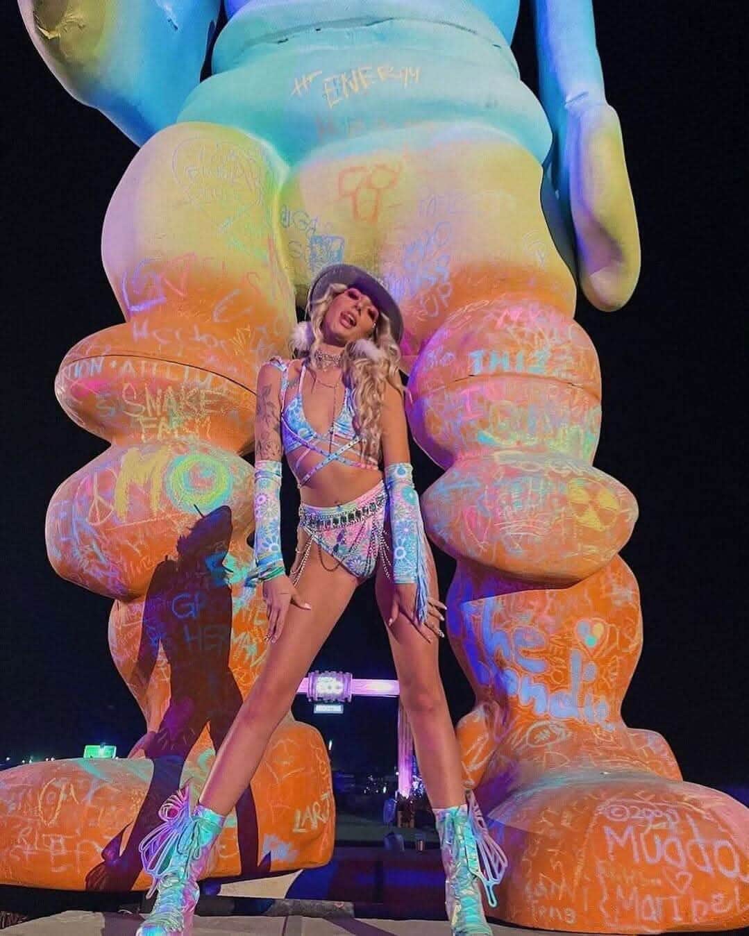 Woman in a pastel wrap bikini top posing in front of a colorful graffiti mural at a vibrant nighttime festival.