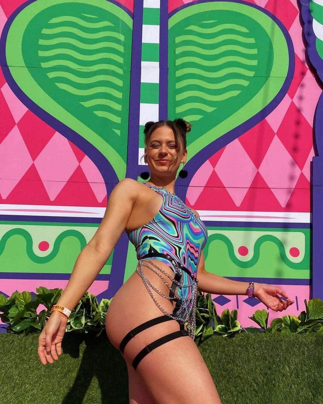 Woman wearing a colorful bodysuit with side chains, posing against a vibrant heart-patterned mural at a rave event.