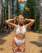Woman in a white rave outfit poses in a forest, showcasing her eco-luxe high-waisted thong and unique accessories.