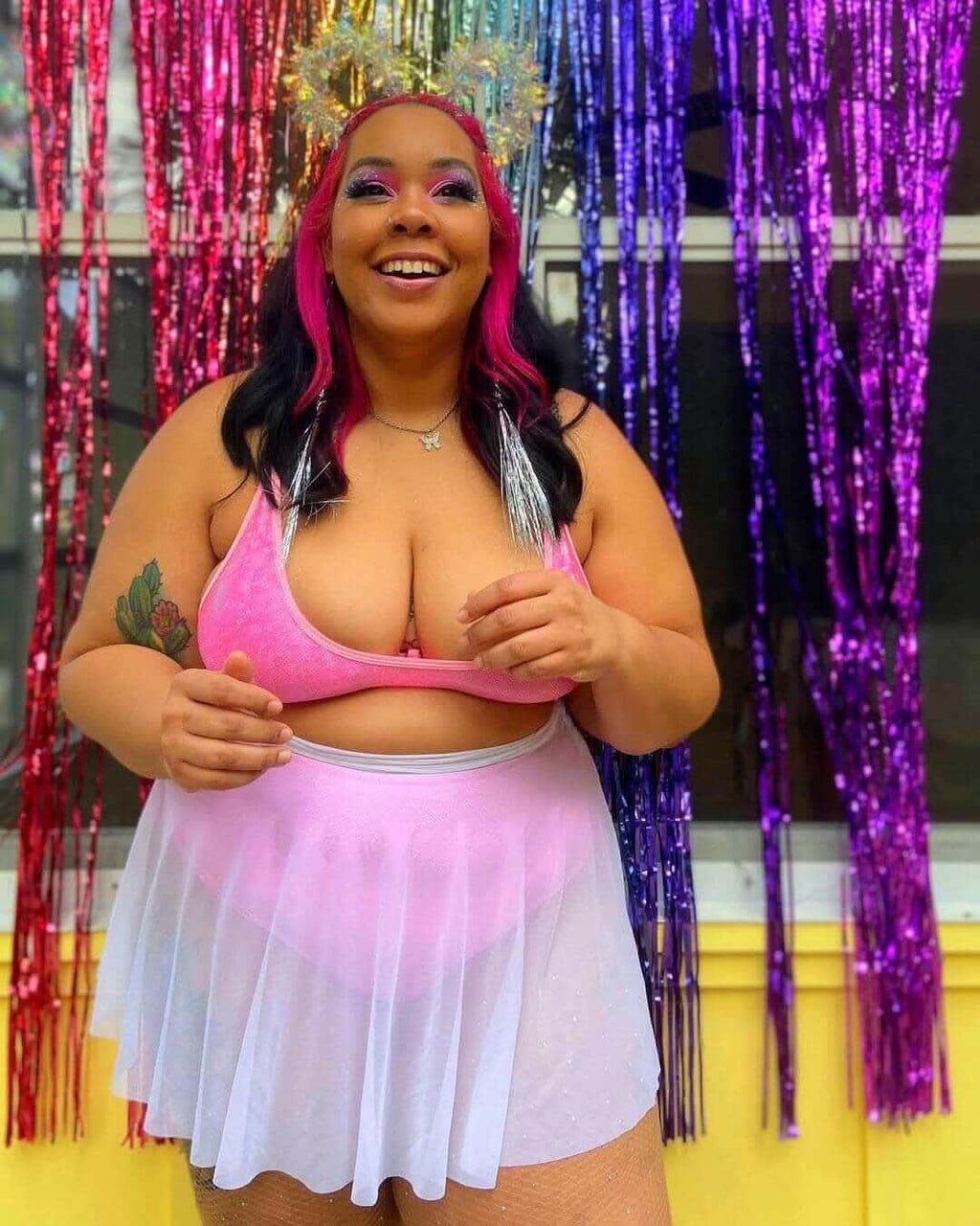 Woman wearing a pink mesh skater skirt and top, smiling joyfully in front of colorful metallic streamers.