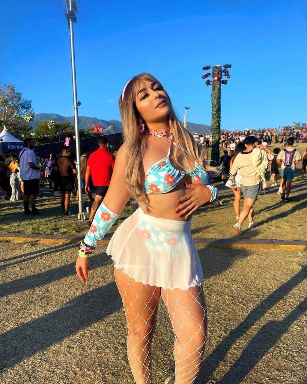 Young woman in a floral crop top and white mesh skater skirt at a lively outdoor festival, surrounded by concert-goers.
